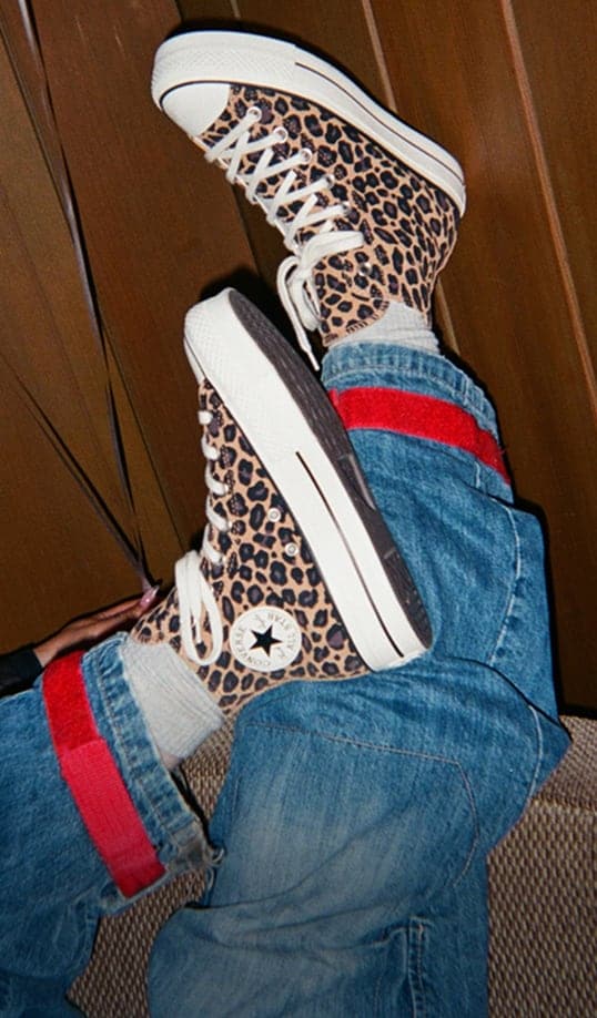 The image shows a pair of legs wearing leopard-print Converse high-top sneakers with white laces and soles. The person is also wearing blue jeans rolled up to reveal gray socks, and the jeans have a distinctive red stripe near the cuffs.