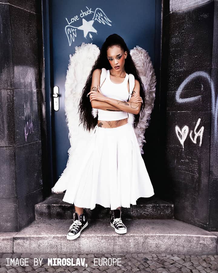 A tattooed woman dressed in white with black sneakers stands confidently with crossed arms in front of a blue door adorned with angel wings and graffiti, creating the illusion that she has wings.