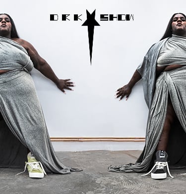 A person stands against a white wall wearing a draped, metallic-silver gown with high slits in a mirrored composition. They wear mismatched sneakers (green on the left, black-and-white on the right) and a stylized black star-logo with text appears above.