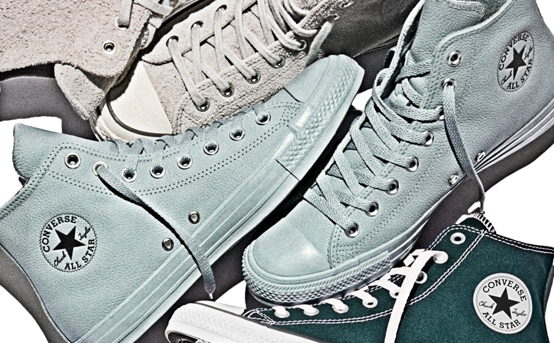 A close-up of several high-top Converse All Star sneakers in shades of teal and grey arranged overlapping. White laces, rubber toe caps, and the circular &ldquo;Converse All Star&rdquo; logo are clearly visible.