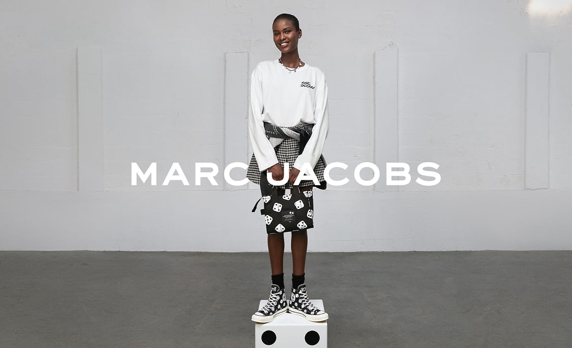A smiling model wearing a white long-sleeve top, patterned skirt, and black-and-white high-top sneakers stands on a white cube against a plain light-gray wall. Bold white text “MARC JACOBS” is overlaid across the center of the image.