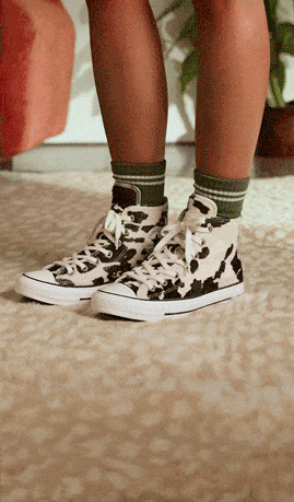 The image shows a pair of legs wearing green striped socks and high-top sneakers with a black-and-white cow print pattern. The person is standing on a textured beige carpet, with part of an orange chair and some greenery visible in the background.