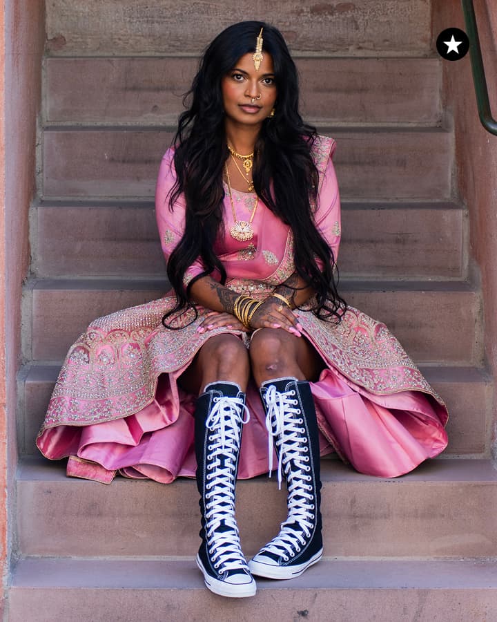 A person dressed in a traditional pink embroidered outfit is sitting on stone steps wearing tall black and white lace-up boots, combining cultural attire with modern footwear.