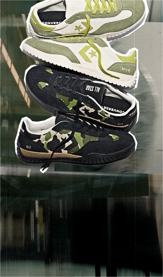 This image shows two pairs of Converse sneakers stacked vertically against a blurred background. The top pair is primarily white with olive green accents, including the laces, side stripe, and heel area. The word &ldquo;MVP&rdquo; is visible on the heel of this pair. The bottom pair features a black base with camouflage patterns in shades of green and beige on the sides. This pair has black laces and displays the text &ldquo;ALL STAR&rdquo; on the tongue and &ldquo;DJ&rdquo; near the heel. Both pairs have a sporty design with textured soles suitable for casual or athletic wear. The overall style suggests a modern, trendy look combining classic Converse branding with unique colorways and patterns.