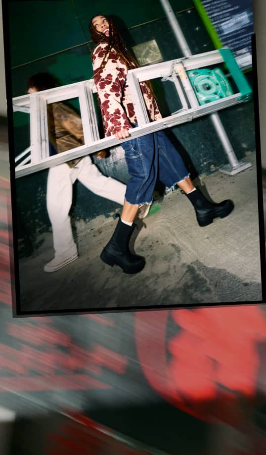 The image shows a person wearing a red and white patterned top, cropped denim pants, black boots, and white socks carrying a metal ladder. Another person dressed in light-colored pants and shoes walks beside them against an industrial background.