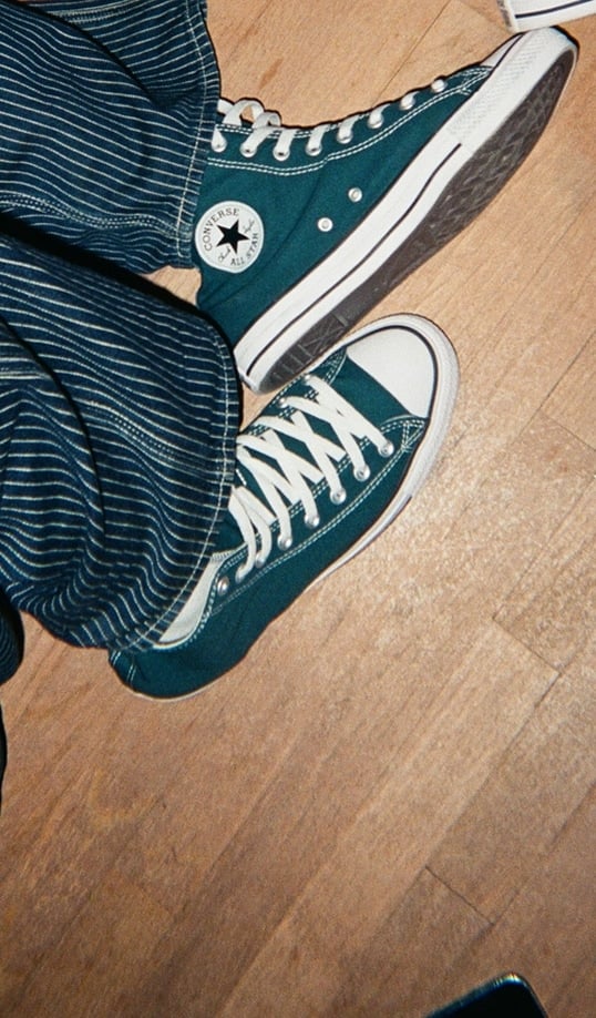 The image shows a person wearing teal Converse All Star high-top sneakers paired with blue and white striped pants. The person is standing on a wooden floor, and the photo is taken from above.