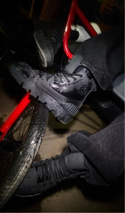A close-up shot of someone wearing black chunky lace-up boots with textured soles, resting their feet on a red bicycle frame and wheel. The scene is dimly lit with shiny, slightly wet surfaces and dark clothing.