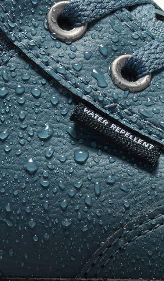 The close-up image shows a blue shoe with water droplets beading on its surface, demonstrating its water-repellent quality. A black tag stitched onto the shoe clearly reads &ldquo;WATER REPELLENT.&rdquo;