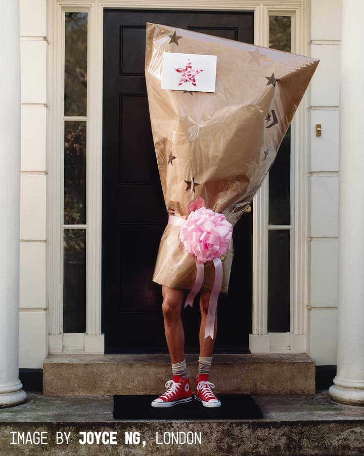A person is standing in front of a black door, completely wrapped in brown star-patterned wrapping paper with a large pink bow around the middle, wearing red high-top sneakers and striped socks.
