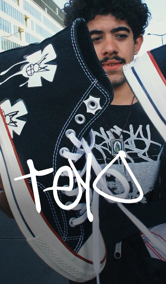 The image shows a person holding up a black high-top sneaker with white laces and a white sole that has red and blue stripes. The sneaker features distinctive white graphic designs resembling stylized flowers or crosses on the side near the ankle area. The person is wearing a black shirt with white graffiti-style text, partially visible behind the shoe, and a necklace with a rectangular pendant featuring an image of what appears to be a religious figure or icon. The person&rsquo;s face is blurred out. There is also large white handwritten text overlaid on the image that reads &ldquo;toyo.&rdquo; In the background, there are modern buildings and street elements indicating an urban setting.