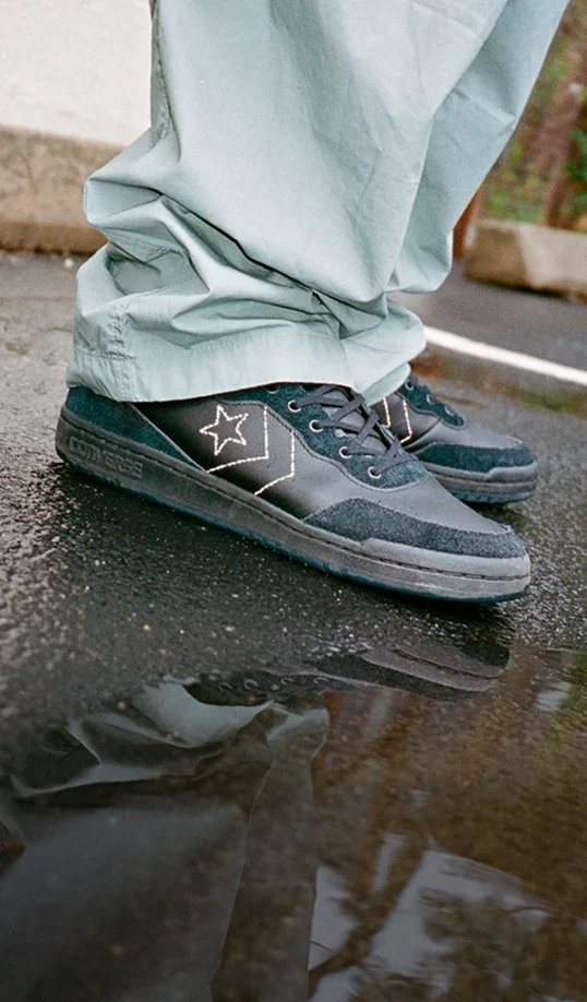 A close-up of black-and-teal Converse-style sneakers worn with light green pants. The shoes stand on wet pavement with a clear reflection visible in a puddle.