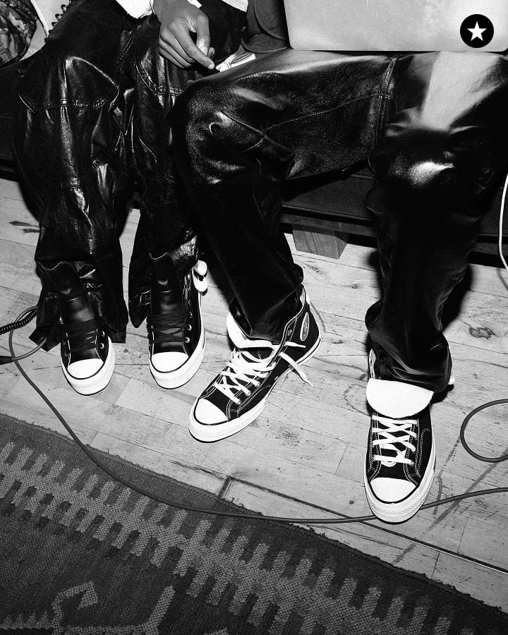 The black-and-white image shows two people sitting side by side, both wearing shiny leather pants and Converse-style sneakers. The focus is on their legs and feet, with a wooden floor and part of a patterned rug visible beneath them.