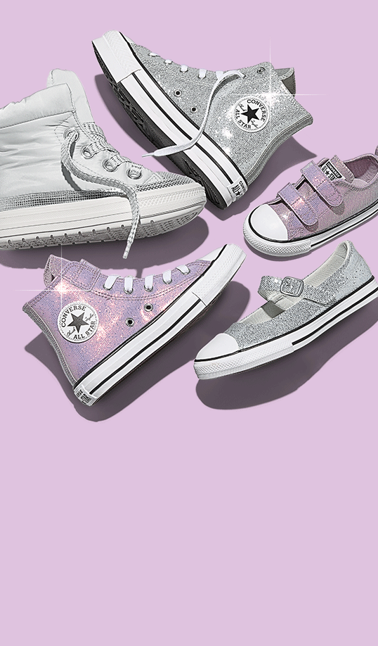 A flat lay of five glittery Converse sneakers in silver and iridescent pink arranged on a lavender background. The shoes include high-tops, low-tops, a Mary Jane strap style, and a children’s Velcro sneaker, all sparkling under studio lighting.