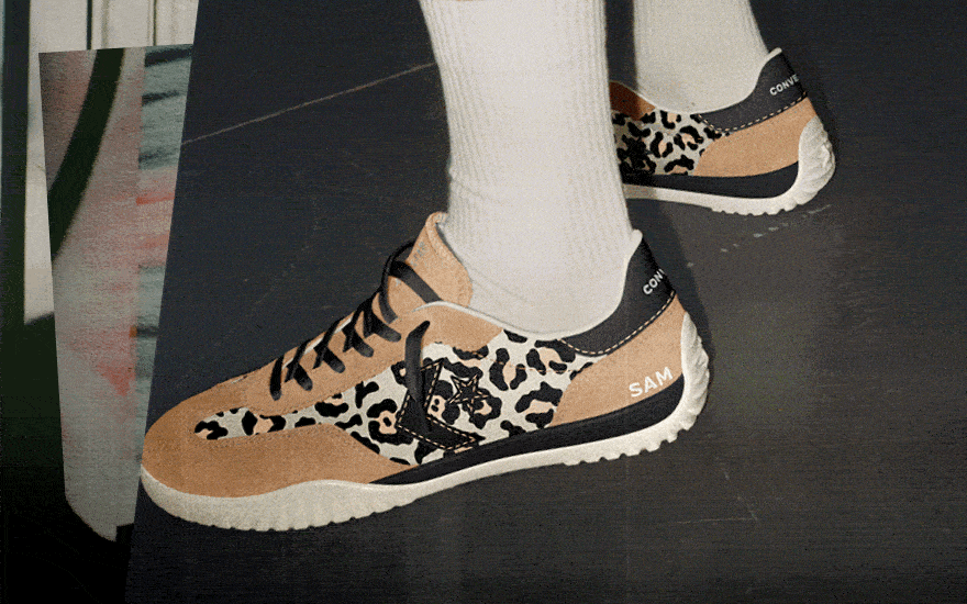 The sneakers feature a tan suede and leopard print design with black laces and white soles. The heel displays the branding &ldquo;CONVERSE&rdquo; and the name &ldquo;SAM.&rdquo;