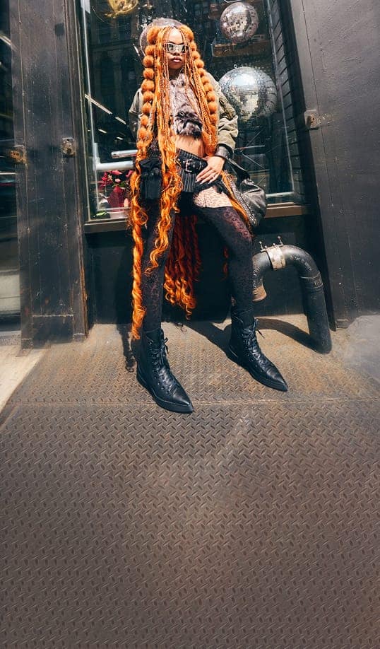 The person stands confidently in front of a dark storefront, showcasing extremely long, bright orange braided hair and wearing black lace leggings with black boots. The background features reflective disco balls inside the window and an industrial pipe to the side.