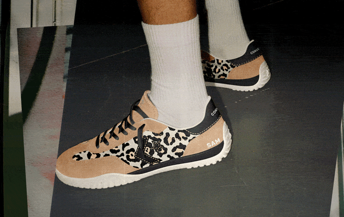 This image shows a close-up of a person&rsquo;s feet wearing sneakers with a distinctive design. The shoes have a beige and black color scheme with leopard print patterns on the sides. The brand &ldquo;CONVERSE&rdquo; is visible on the heel tab, and the name &ldquo;SAM&rdquo; is printed in white near the back of one shoe. The person is also wearing plain white crew socks, and they are standing on a dark floor surface that appears to be indoors. The overall style of the sneakers combines animal print with classic sporty elements.