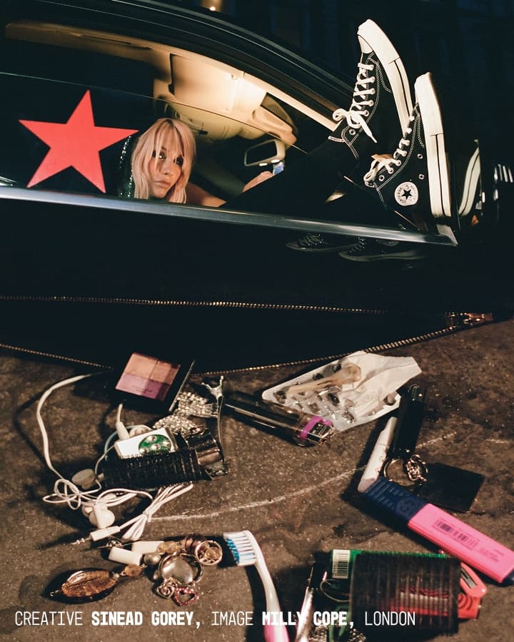 The image depicts a person with blurred face sitting in a black car with their legs out the window wearing black Converse sneakers, while various personal items like makeup, jewelry, earphones, and a toothbrush are scattered on the ground outside.