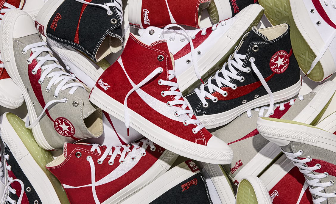 A pile of high-top canvas sneakers in red, black, gray, and white, layered with visible white laces and rubber soles. Several shoes show branded text, including the &ldquo;Converse All Star&rdquo; patch and the &ldquo;Coca‑Cola&rdquo; logo.