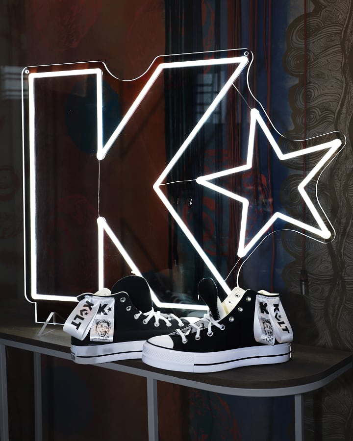The image shows a pair of black and white high-top sneakers displayed on a table in front of a large illuminated neon sign shaped like the letter &ldquo;K&rdquo; with a star. The sneakers have tags attached to them featuring illustrations and the text &ldquo;KLT.&rdquo;