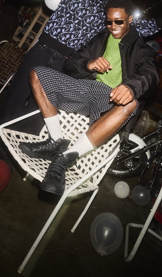 The image shows a person sitting on a chair smiling while wearing black high top Chucks. Overlaid text reads ”Styled Winter. In the shoes that started it all.”