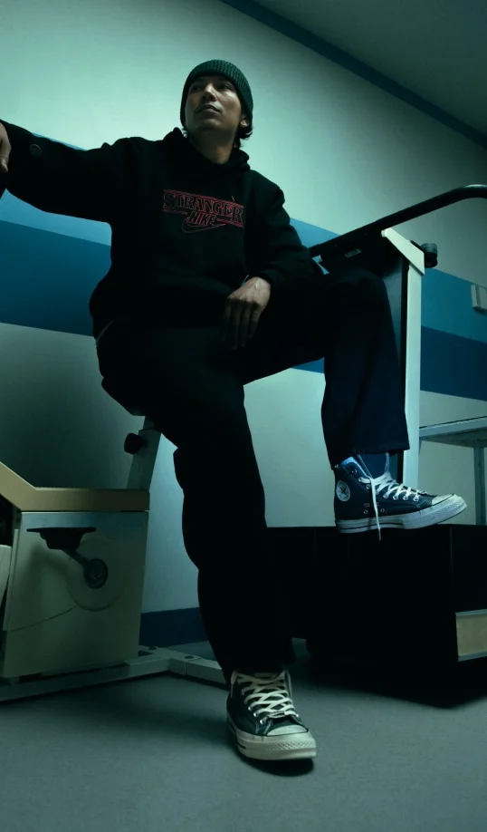A person in a dark beanie and a black hoodie reading “STRANGER” sits with one foot on a raised platform, wearing high-top Converse sneakers and looking upward. The moody teal-lit room shows a striped wall and clinical-looking furniture, including an exam chair and a handrail, in the background.