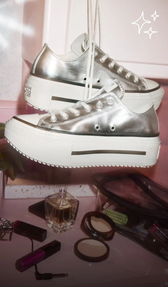 A pair of metallic silver platform sneakers hang by their laces above a cluttered vanity. Below them are makeup items and a perfume bottle scattered on a pink countertop.
