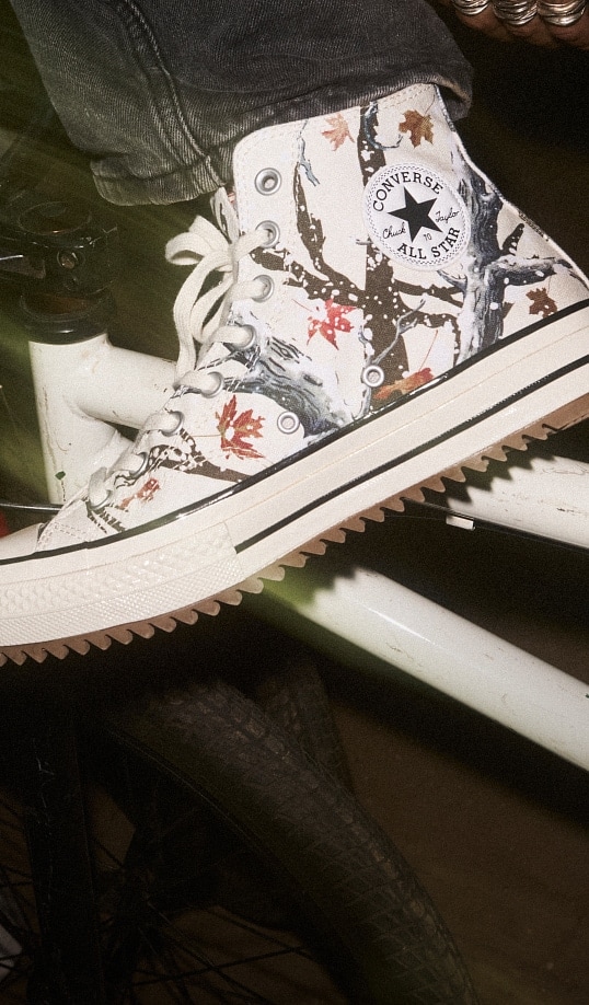 A close-up of a white Converse Chuck Taylor high-top sneaker with a tree-and-autumn-leaf print and the &ldquo;Converse All Star&rdquo; logo visible on the ankle. The shoe, laced and worn with dark jeans, rests on the frame of a bicycle above the tire.