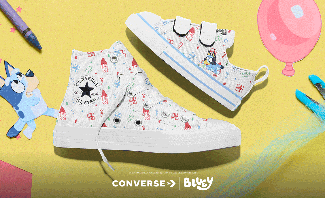 The image shows a pair of Converse shoes featuring a playful Bluey cartoon design on a yellow background with colorful craft items around. The larger shoe is a high-top with laces, and the smaller one is a low-top with velcro straps, both decorated with holiday-themed patterns and Bluey characters.