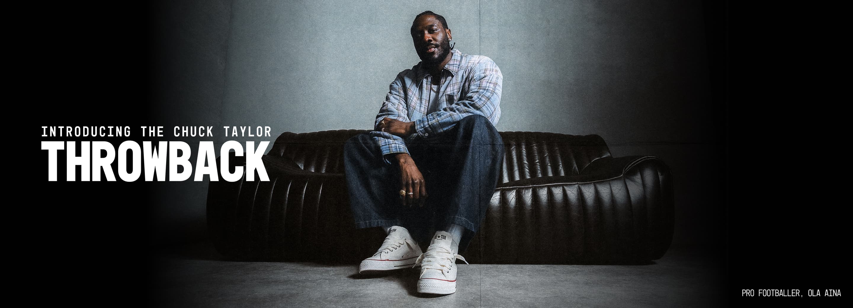 Pro Footballer, Ola Aina, in a plaid shirt and white sneakers sits on a dark leather sofa. The image includes a headline &ldquo;THROWBACK&rdquo; for the White high-top Converse Chuck Taylor sneakers