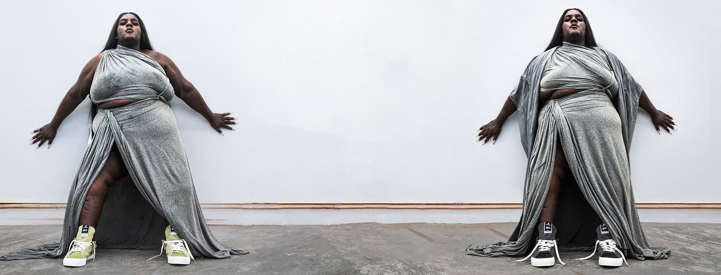 A person stands against a white wall wearing a draped, metallic-silver gown with high slits in a mirrored composition. They wear mismatched sneakers (green on the left, black-and-white on the right) and a stylized black star-logo with text appears above.