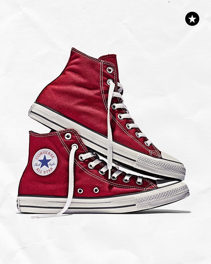 The image shows a pair of red Converse All Star sneakers, with one shoe standing upright displaying the logo patch and the other lying flat showing the laces and insole.