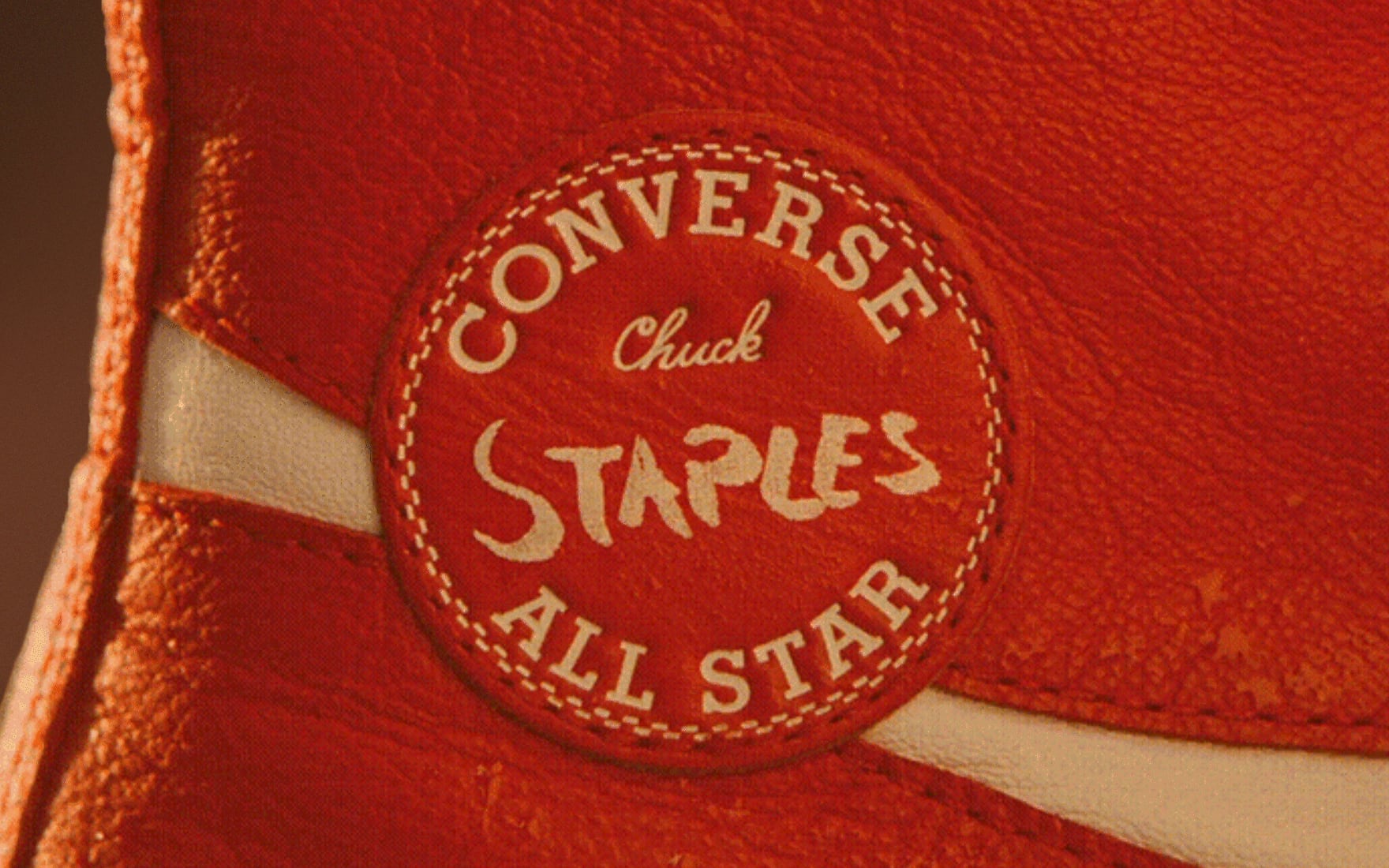 The image shows a close-up of a red Converse Chuck Taylor All Star shoe patch with the word &ldquo;STAPLES&rdquo; prominently written in the center. The patch is stitched onto the leather material of the shoe, highlighting its texture and detail.