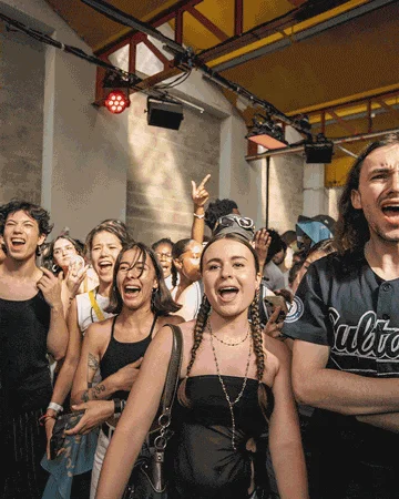 The image shows a group of people gathered indoors under a yellow ceiling with exposed beams and lighting equipment. Most individuals are casually dressed, some making hand gestures, suggesting a social or party atmosphere.