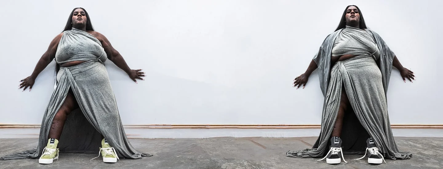 A person stands against a white wall wearing a draped, metallic-silver gown with high slits in a mirrored composition. They wear mismatched sneakers (green on the left, black-and-white on the right) and a stylized black star-logo with text appears above.