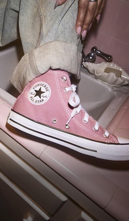 A person wearing a pink Converse All Star high-top sneaker rests their foot on a pink-tiled bathroom sink edge. A hand with long, manicured nails and a ring hangs nearby above the shoe.