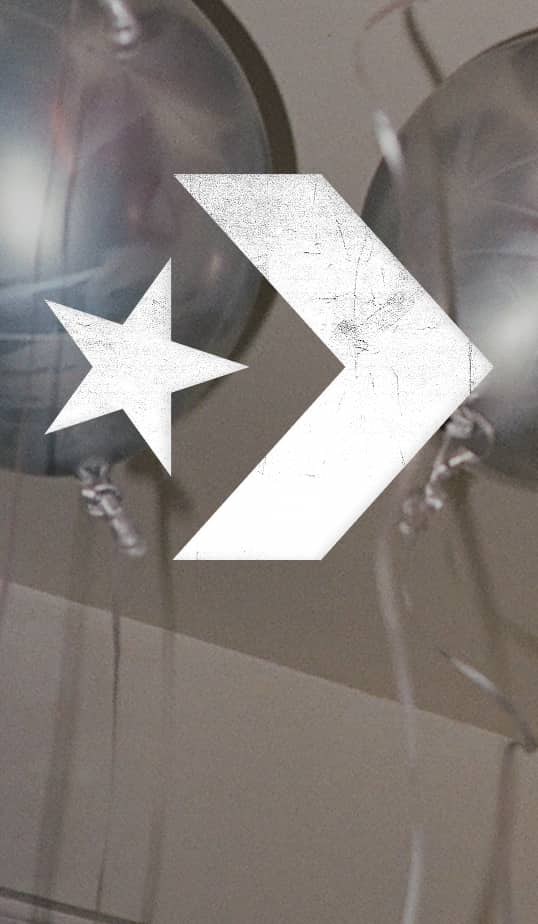 A large white, distressed chevron-and-star graphic is overlaid in the center of the image. Behind it are out-of-focus metallic gray balloons with visible strings hanging downward.