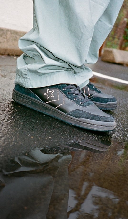 A close-up of black-and-teal Converse-style sneakers worn with light green pants. The shoes stand on wet pavement with a clear reflection visible in a puddle.