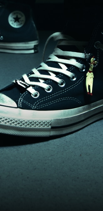 A close-up of a black high-top canvas sneaker with white laces and a thick rubber sole. A small charm shaped like a yellow chicken hangs from the shoe’s eyelets, while another sneaker with a visible star logo sits blurred in the background.