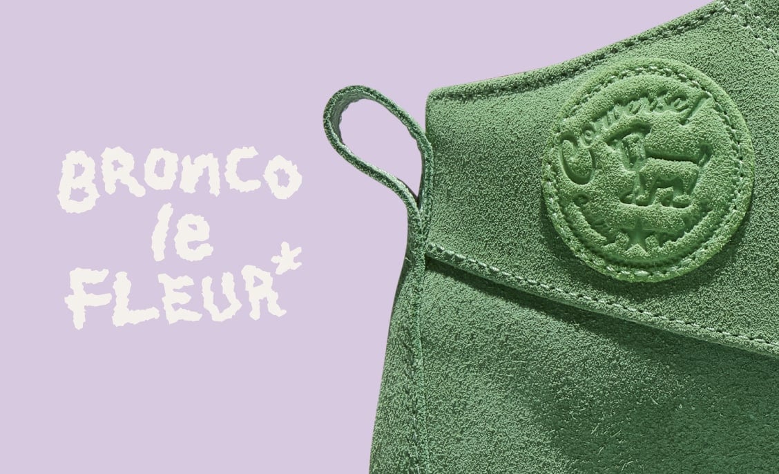 A close-up of a green suede shoe ankle with a loop and an embossed circular Converse-style logo on a lavender background. To the left, hand-drawn white text reads 