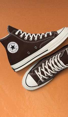 The image shows a pair of dark brown high-top Converse All Star sneakers with white laces and rubber soles on an orange background. One shoe displays the iconic circular Converse logo patch, while the other is shown from a top-down view.