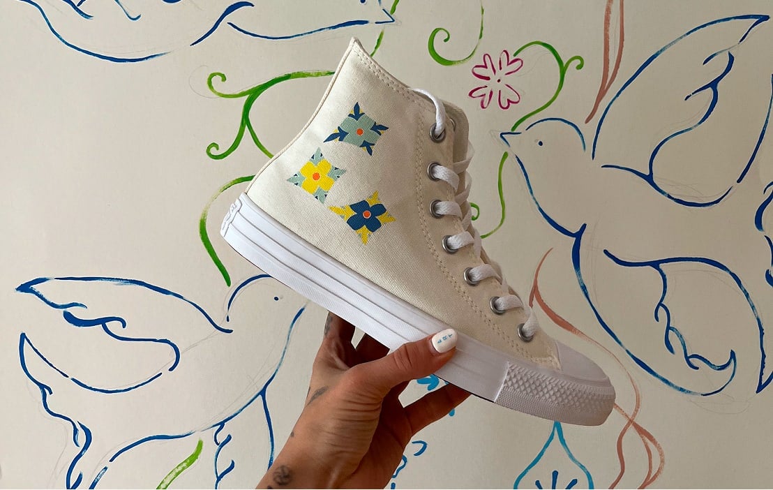 The image shows a hand holding up a beige high-top sneaker with white laces and a white rubber sole. The sneaker has three colorful flower-like designs on the side in blue, yellow, and green colors. The background features a wall painted with simple, stylized blue birds outlined in blue lines along with some green vines and small pink flowers. The overall scene has an artistic and casual vibe, combining the shoe&rsquo;s design with the mural behind it. The person&rsquo;s nails are painted white with small blue details visible on one nail.