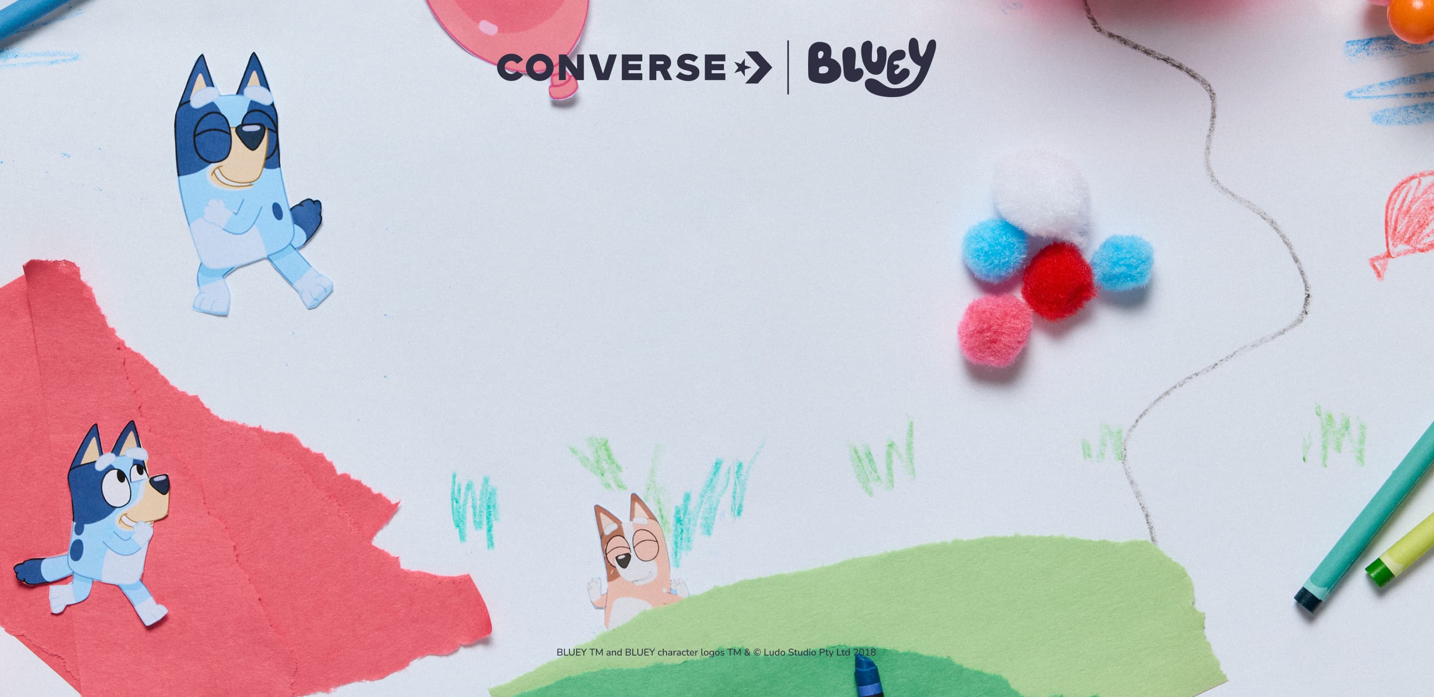 This image features paper cutouts of Bluey characters on torn red and green paper, with crayon drawings of grass and balloons on a white background. Colorful pom-poms arranged like flowers and two crayons add playful, crafty details around the &ldquo;CONVERSE | BLUEY&rdquo; logo at the top center.