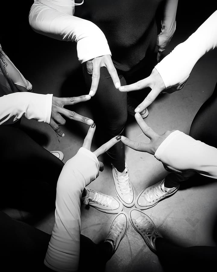 Five people wearing white long-sleeve tops and white sneakers stand in a circle with their feet together while forming a star shape with their fingers above.