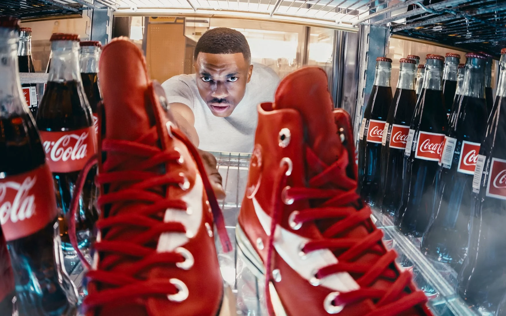 The image shows a person reaching into a refrigerator stocked with glass bottles of Coca-Cola and a pair of red high-top sneakers placed inside. The perspective is from inside the fridge looking outward toward the person.