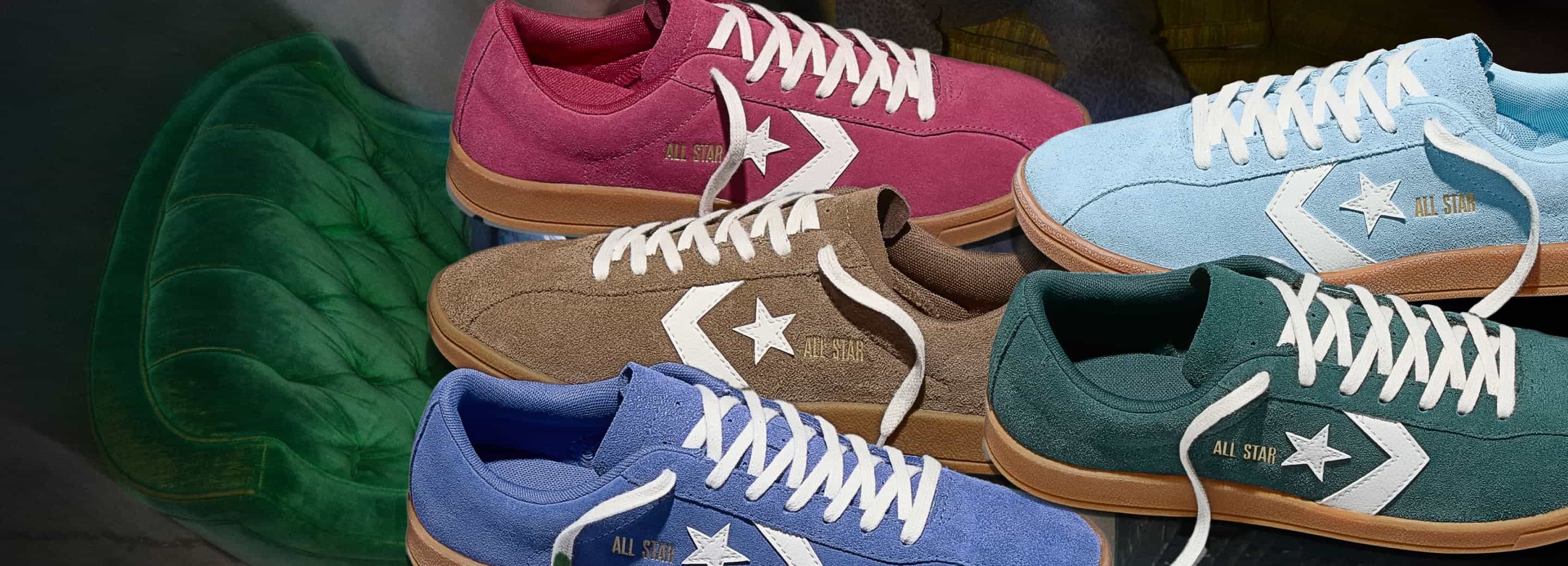 Several pairs of suede sneakers in bright colors&mdash;pink, brown, light blue, royal blue, and dark green&mdash;are arranged overlapping, each with white laces, a white star chevron logo, and gum-colored soles. They rest on or above a deep green, tufted velvet surface.