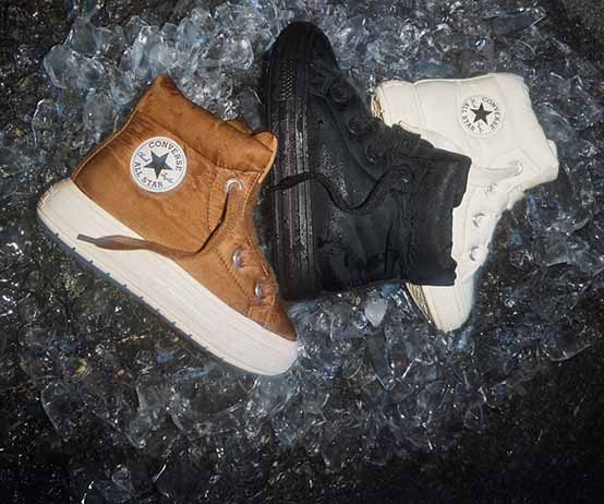 Three high-top Converse boots&mdash;brown, black, and white&mdash;are arranged together on a bed of crushed ice. Each boot shows the circular &ldquo;Converse All Star&rdquo; logo on the ankle panel.