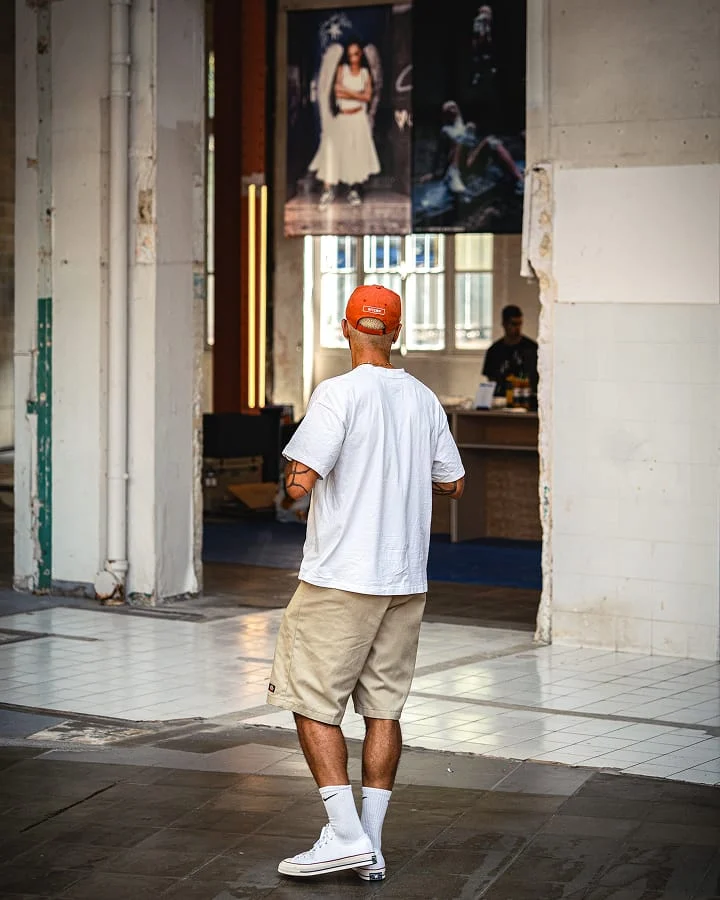 The image shows a man standing indoors with his back to the camera, wearing a white t-shirt, beige shorts, white socks, white sneakers, and an orange cap. In the background, there are two large posters featuring people and a person behind a counter in a well-lit room.