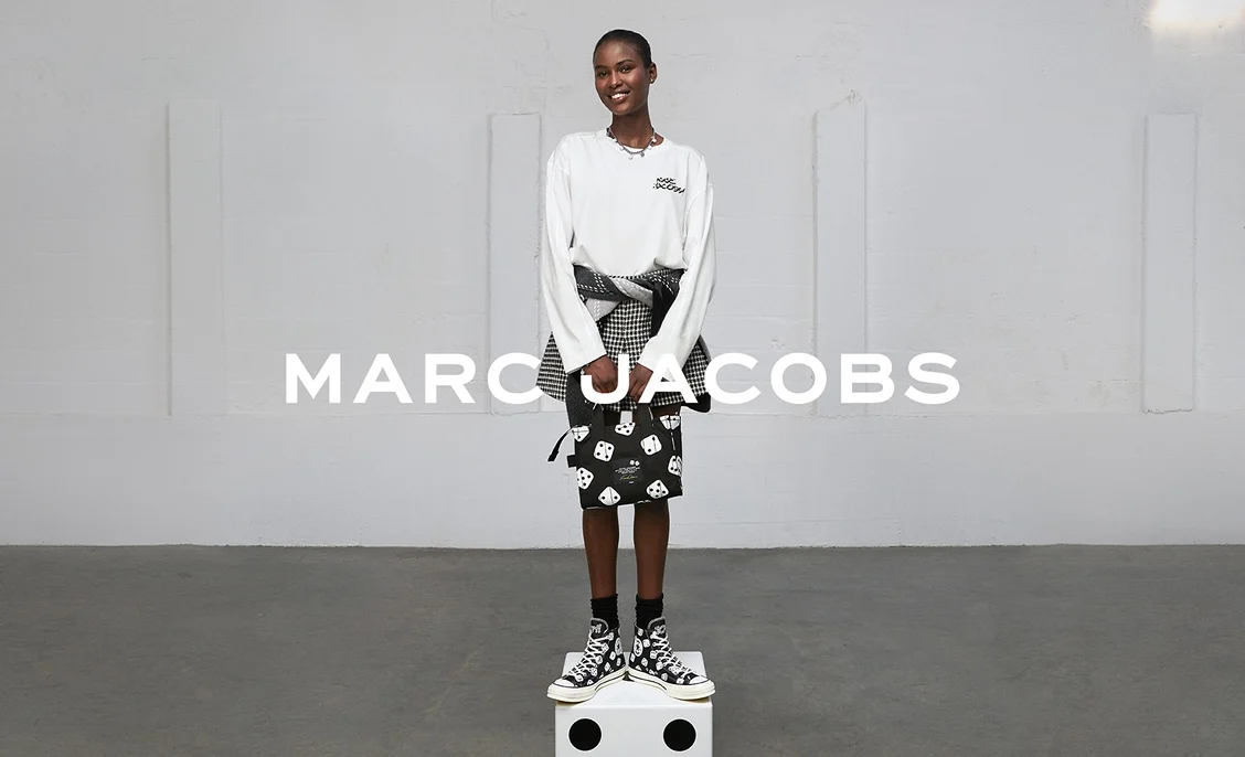 A smiling model wearing a white long-sleeve top, patterned skirt, and black-and-white high-top sneakers stands on a white cube against a plain light-gray wall. Bold white text &ldquo;MARC JACOBS&rdquo; is overlaid across the center of the image.