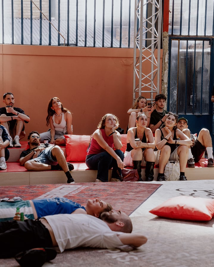 The image depicts a group of people sitting on tiered seating against an orange wall, with two individuals lying on the floor in the foreground. The setting appears casual and relaxed, with some people holding drinks and others using their phones.