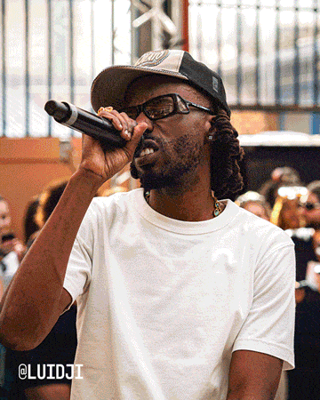 The image shows a person wearing a white t-shirt and a cap, holding a microphone and seemingly performing or speaking in front of an audience. The background includes several people, some with blurred faces, and the text “@LUIDJI” is visible in the bottom left corner.